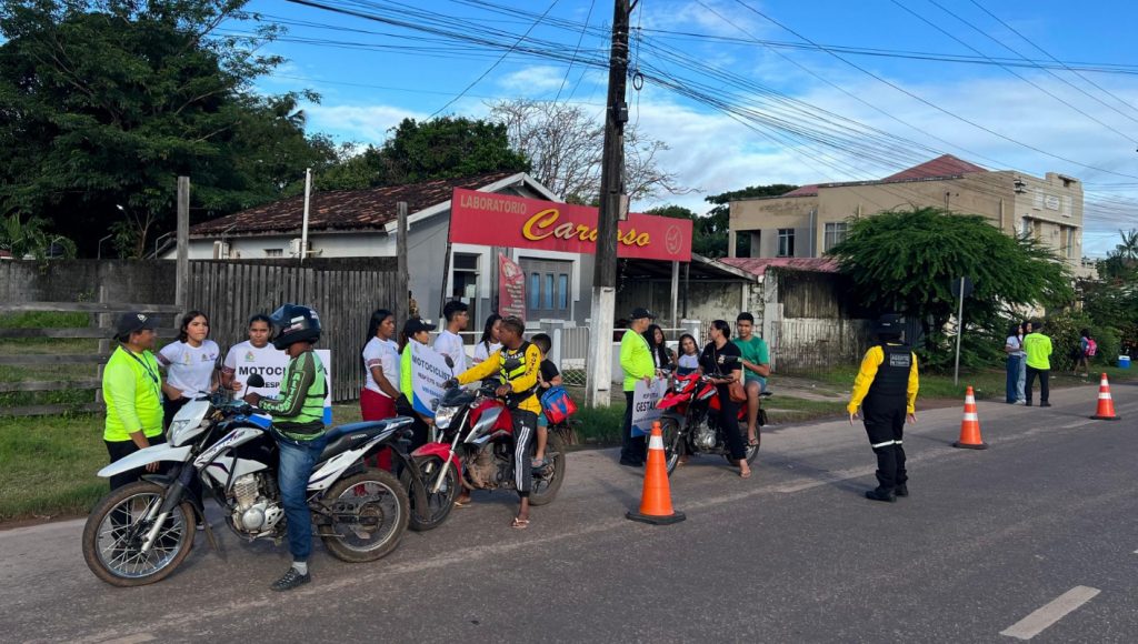 Fotografia registra o momento que um blitz é realizada. Na foto, há várias pessoas com uniformes de agente de trânsito, estudantes com uniformes escolares e condutores de motocicletas em suas motocicletas. Há cones sinalizando a rua.